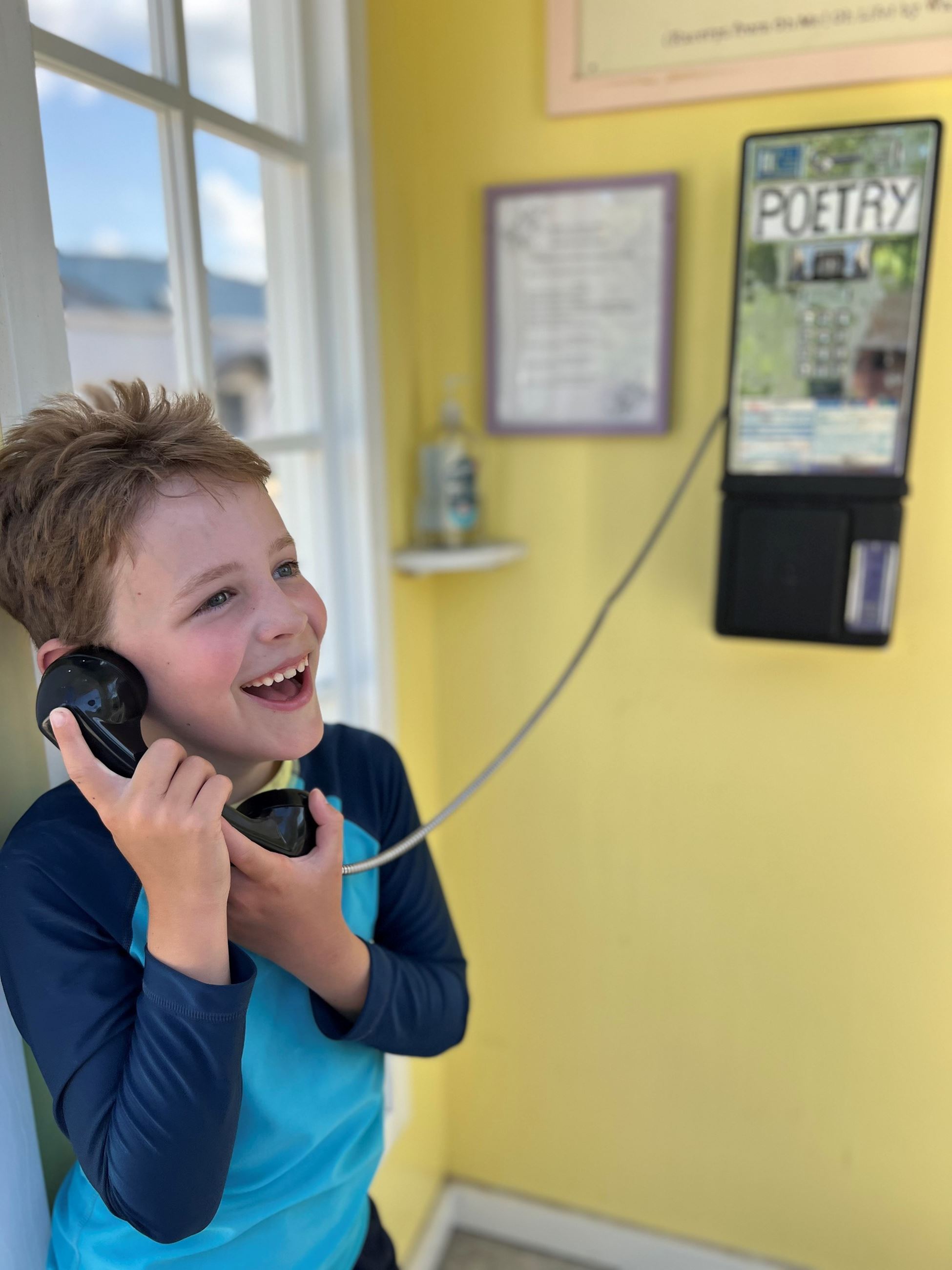 Boy in a black and blue shirt, smiles while listening to a pay phone